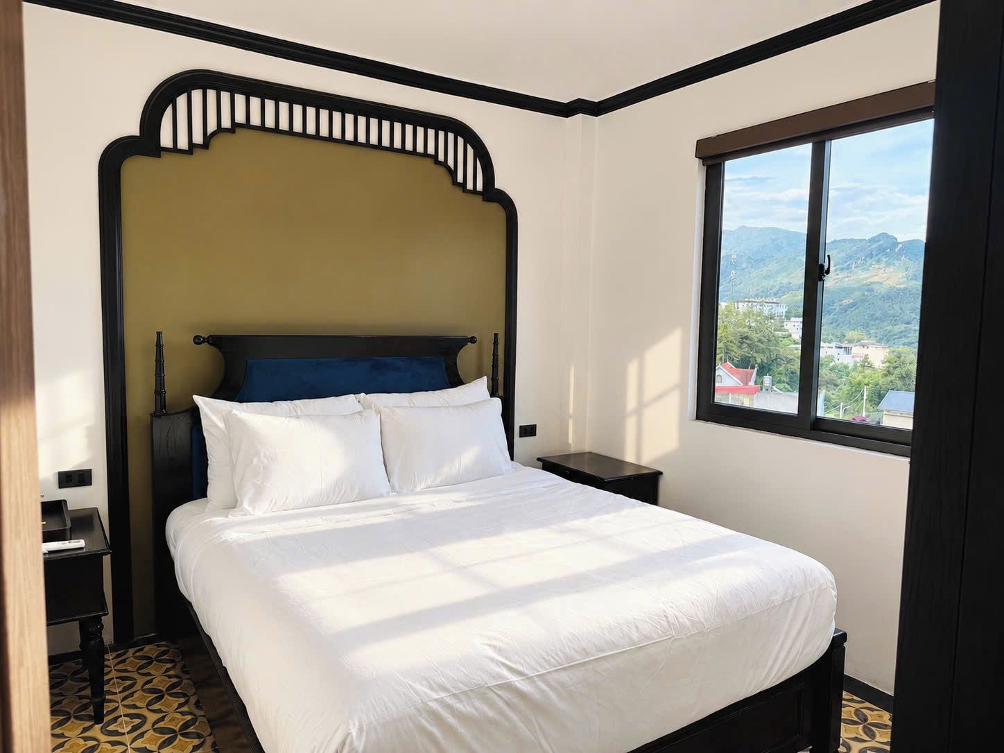 Habitación de hotel con cama, mesita de noche, ventana con vistas y suelo estampado.