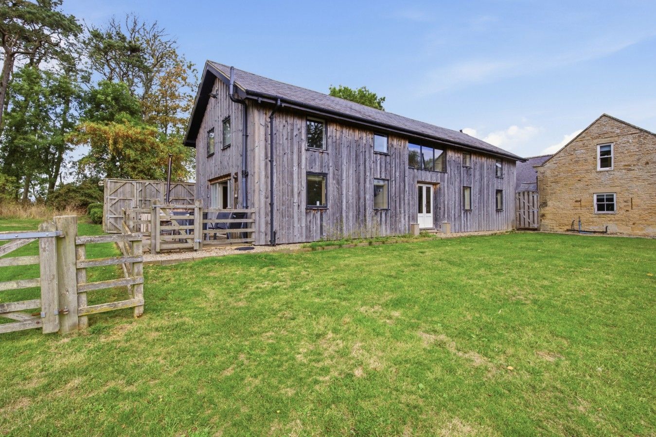 A rustic barn with a stone building adjacent to it, surrounded by a grassy field and a wooden fence.