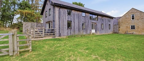 Une grange rustique, adossée à un bâtiment en pierre, entourée d’un champ herbeux et d’une clôture en bois.