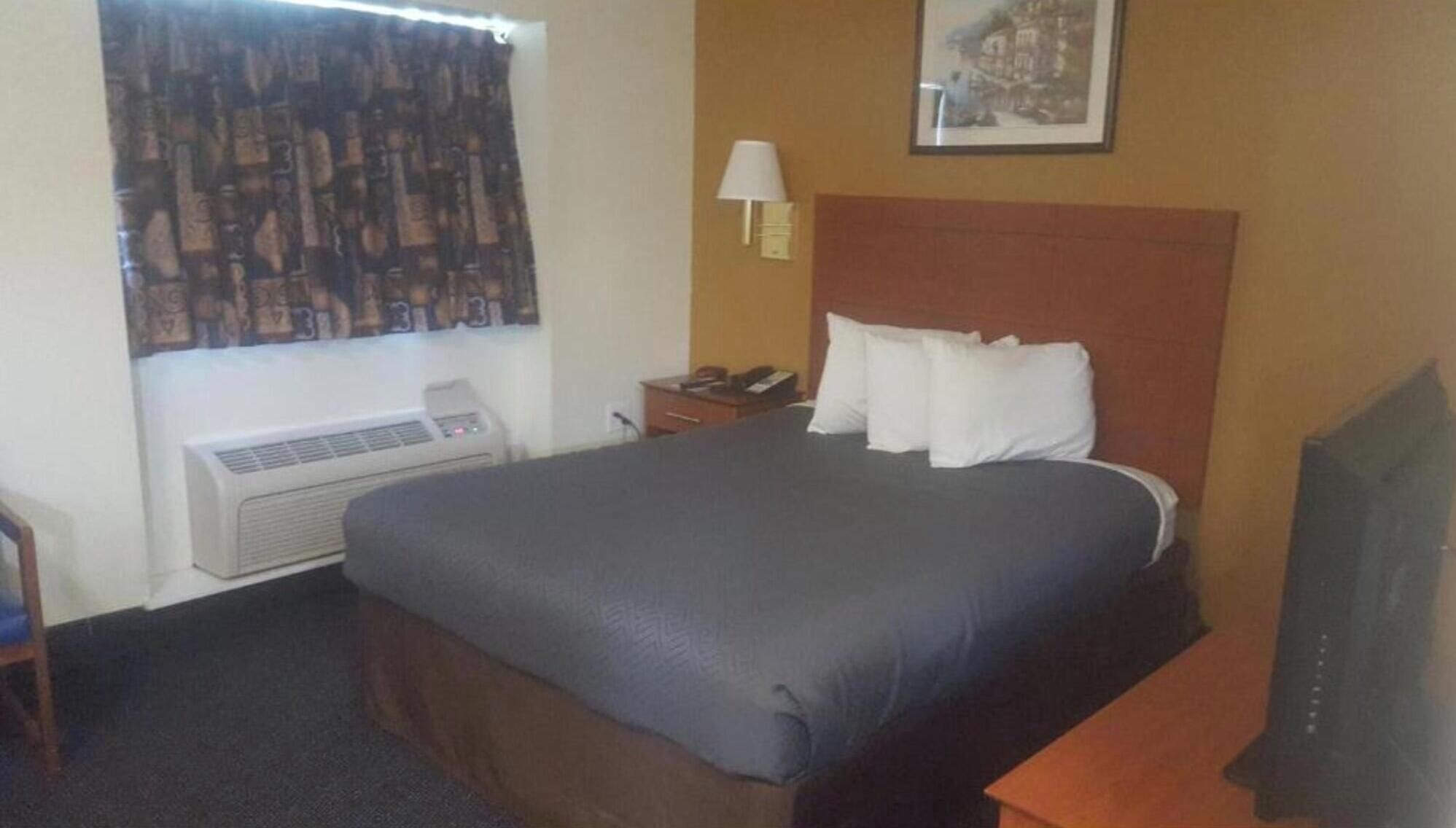 Habitación de hotel con cama, mesita de noche, lámpara, ventana con cortinas e imagen colgada en la pared.