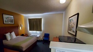 A hotel room with a bed, a desk, a chair, a window with curtains, and a wall-mounted lamp. - 1 Queen Bed | Smoking, Kitchen (Richmond VA)