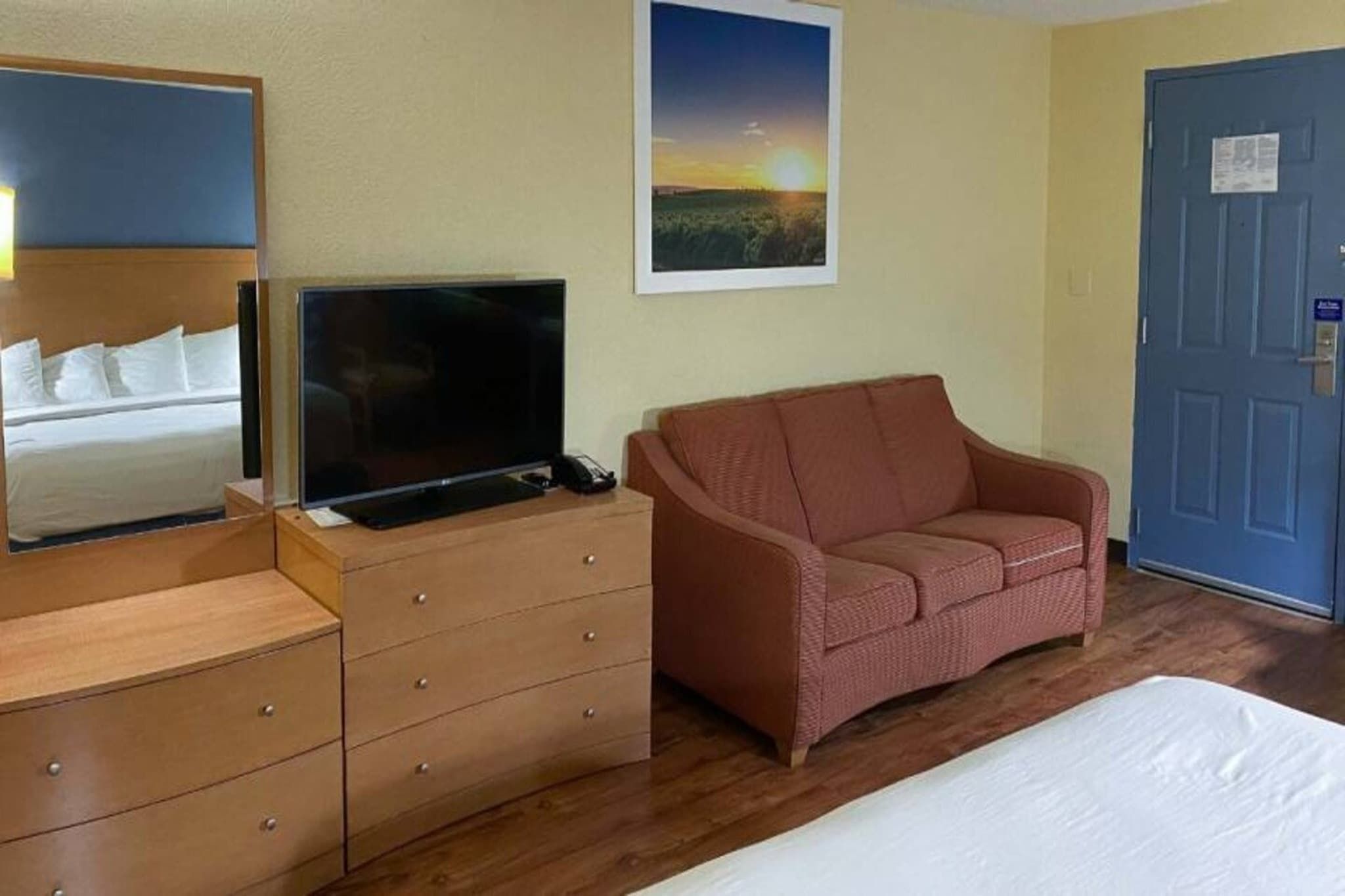 A hotel room with a bed, a television on a wooden dresser, a sofa, a painting on the wall, and a door.