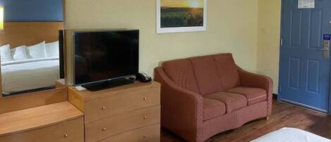 A hotel room with a bed, a television on a wooden dresser, a sofa, a painting on the wall, and a door.
