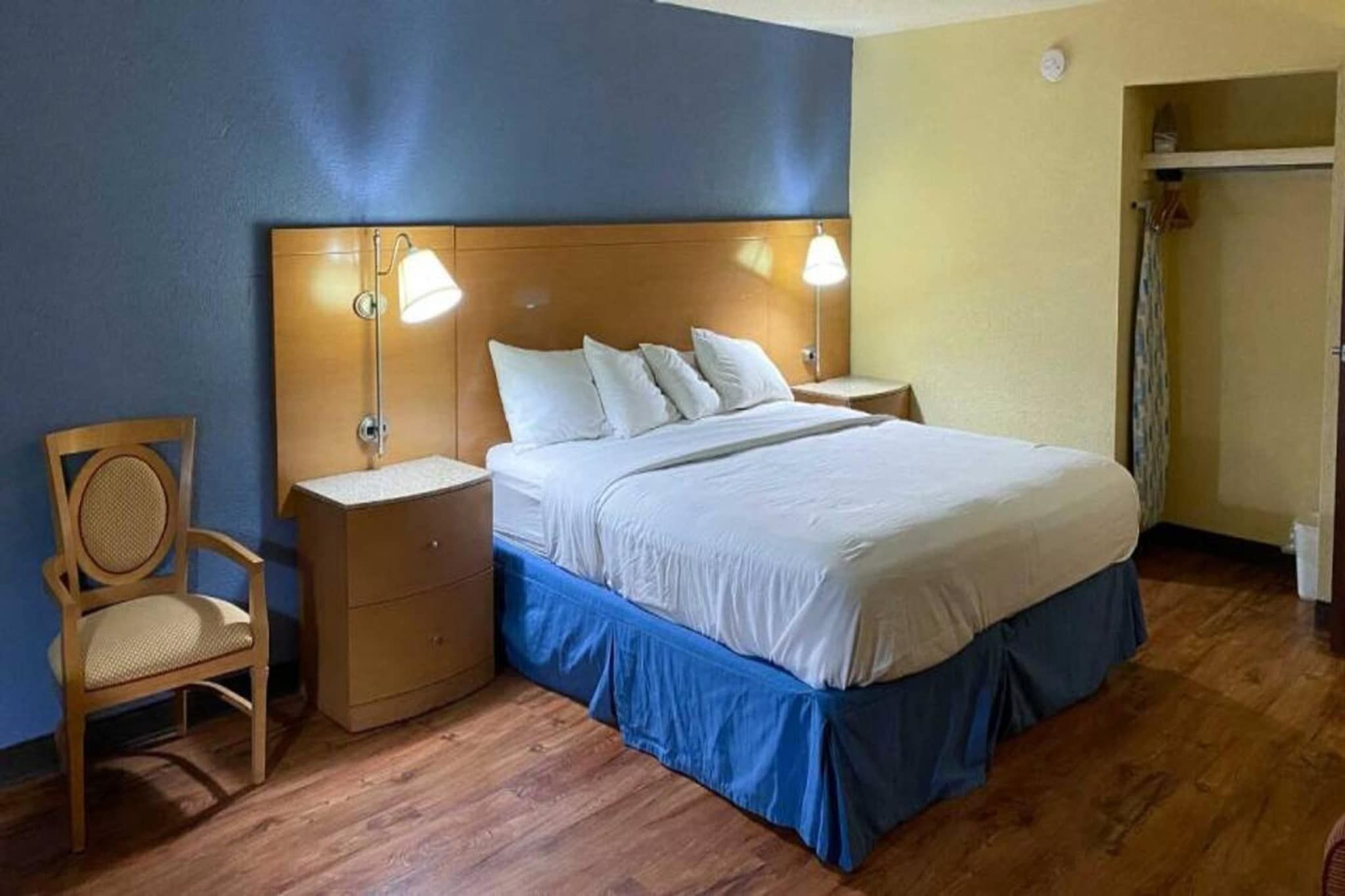 A hotel room with a wooden headboard, a single bed with white bedding, a nightstand with a lamp, a wooden chair, and a wooden floor.