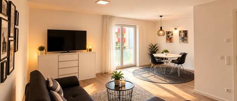 A modern living room with a sofa, a flat-screen TV, a dining area with a table and chairs, and a sliding glass door leading to an outdoor balcony.