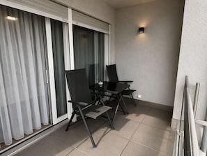 A balcony with a glass table and two chairs, a sliding glass door, and a textured wall.