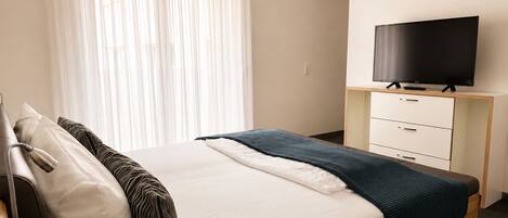 A hotel room with a bed, a television on a stand, a bedside table, and a window with curtains.