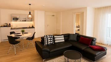 Ein modernes Wohnzimmer mit einem schwarzen Ecksofa, einem runden Couchtisch und einem Essbereich mit weißem Tisch und schwarzen Stühlen.