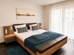 A modern bedroom with a large bed, a bedside table, a nightstand, a wall-mounted lamp, and a cityscape painting.