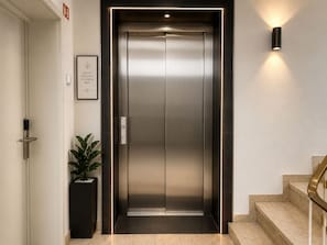 A modern elevator with a metallic finish, a framed sign above it, and a potted plant beside the elevator.