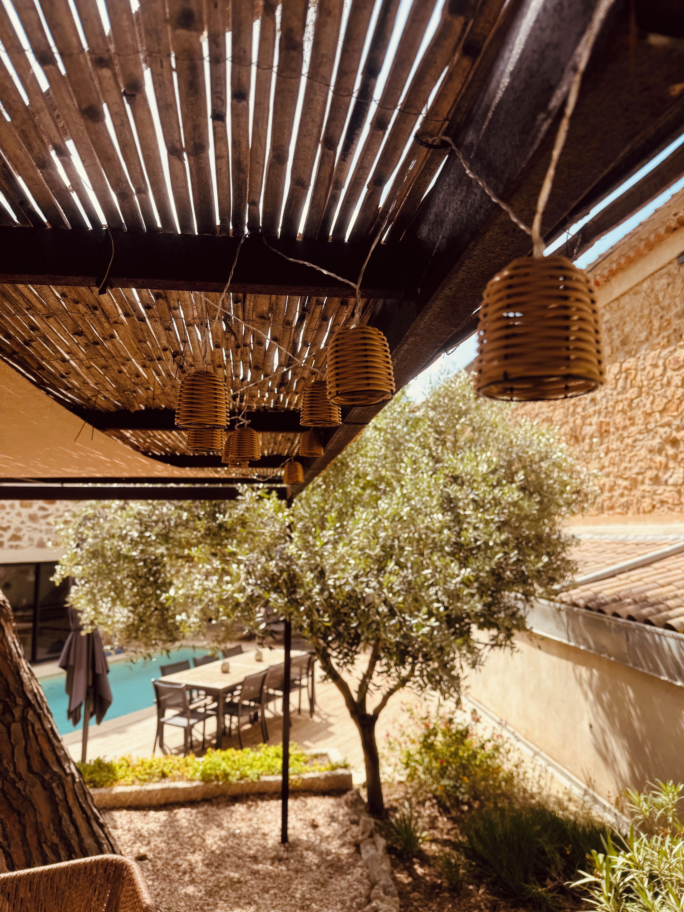 A covered outdoor area with a wooden ceiling, hanging wicker lamps, a table with chairs, and a pool in the background.