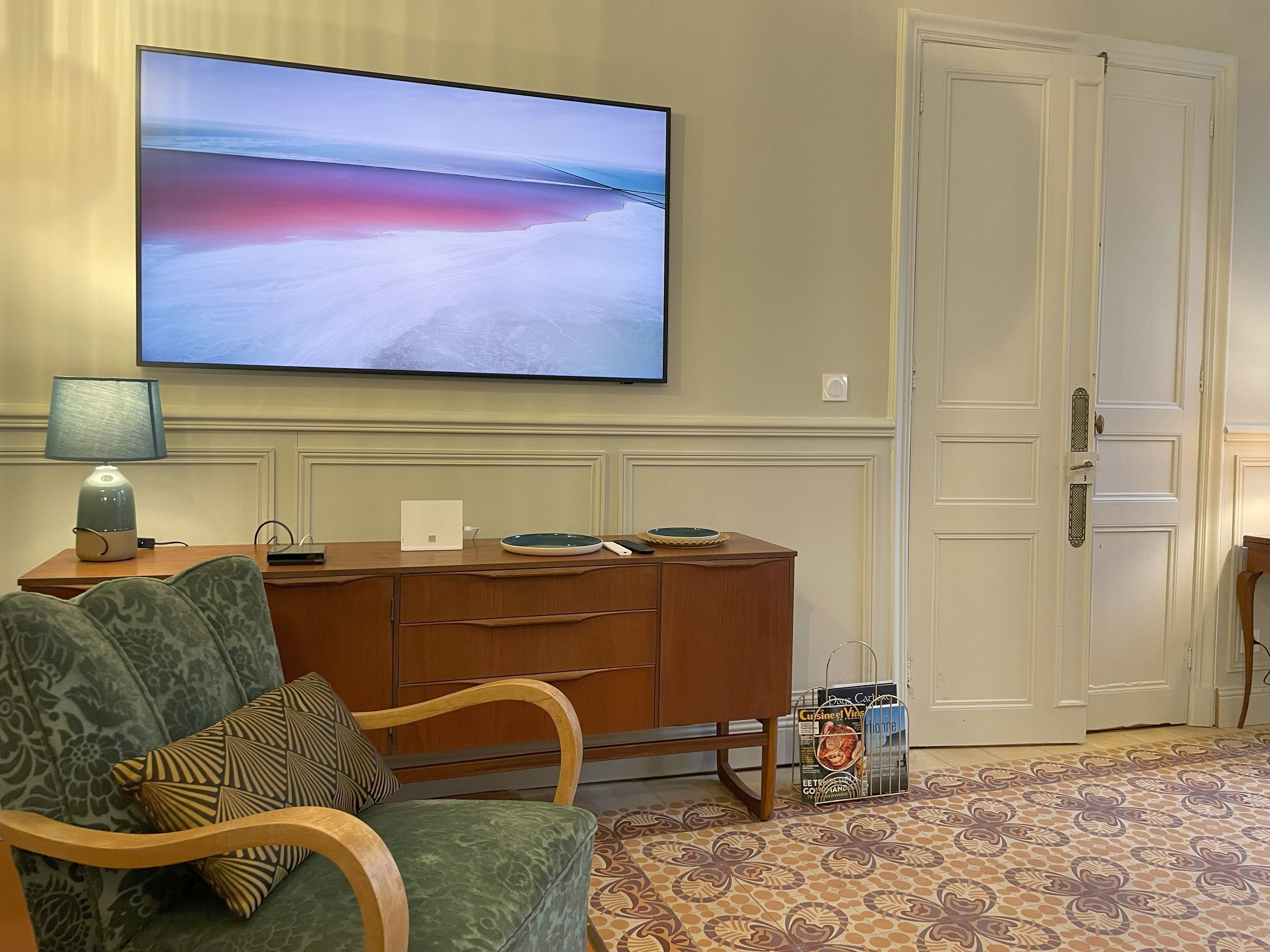 A living room with a flat-screen TV mounted on the wall, a wooden sideboard, a green sofa with patterned cushions, and a patterned carpet.