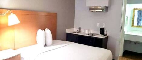Habitación de hotel con cama, mesita de noche, lámpara y una pequeña cocina con lavabo y encimera.