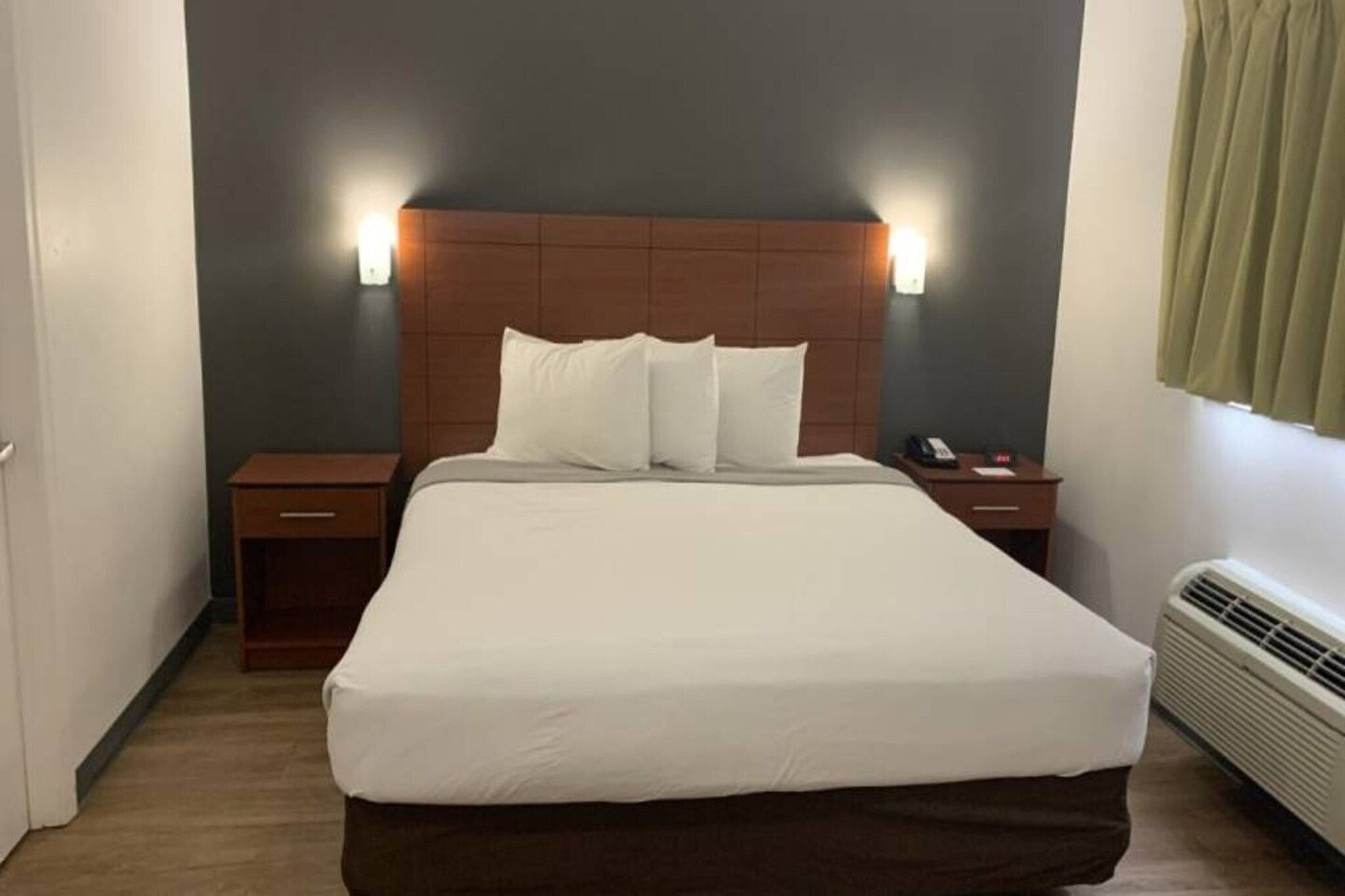 Una habitación de hotel con una cama grande, dos mesitas de noche, una lámpara de pared y un ventanal con cortinas.