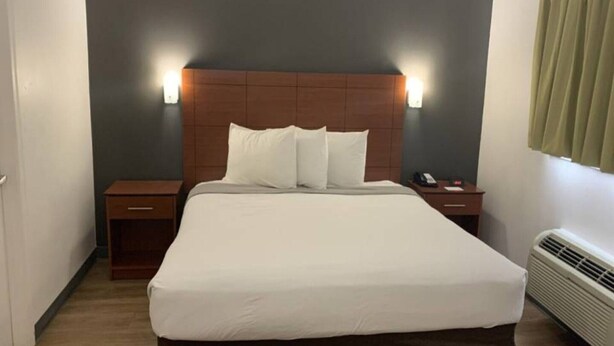 Et hotelværelse med en stor seng, to sengeborde, en lampe på væggen og et vindue med gardiner.