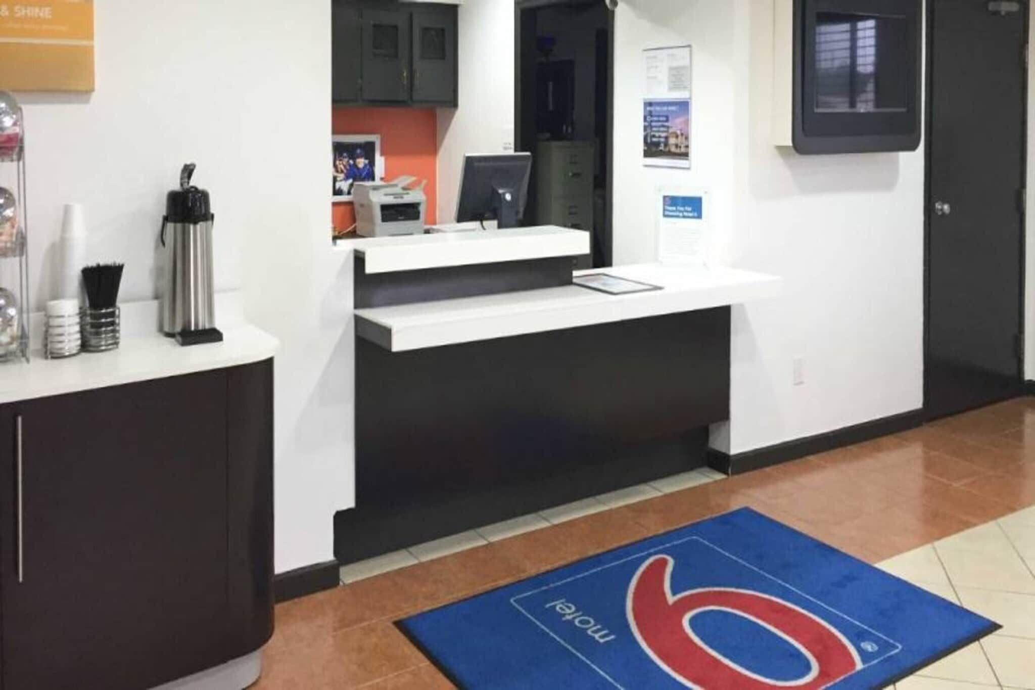A reception desk with a computer, a telephone, and a coffee machine. There is a blue mat with a logo on the floor.