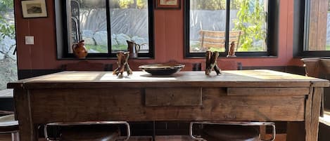 A rustic kitchen with a large wooden table, two stools, and a window with a view of greenery.