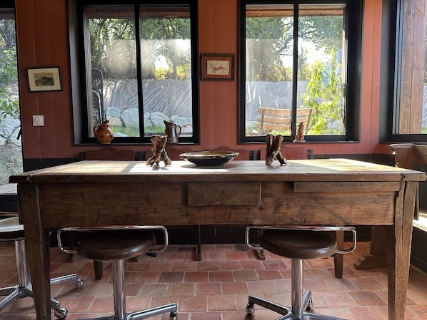 A rustic kitchen with a large wooden table, two stools, and a window with a view of greenery.