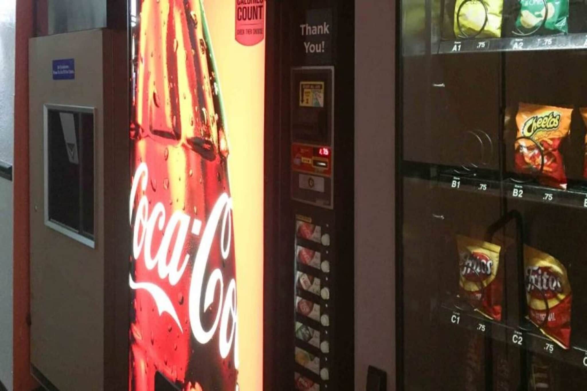 Un distributeur automatique orné du logo Coca-Cola, d’une pancarte indiquant « Merci ! » et proposant des en-cas comme des Cheetos et des Pringles.