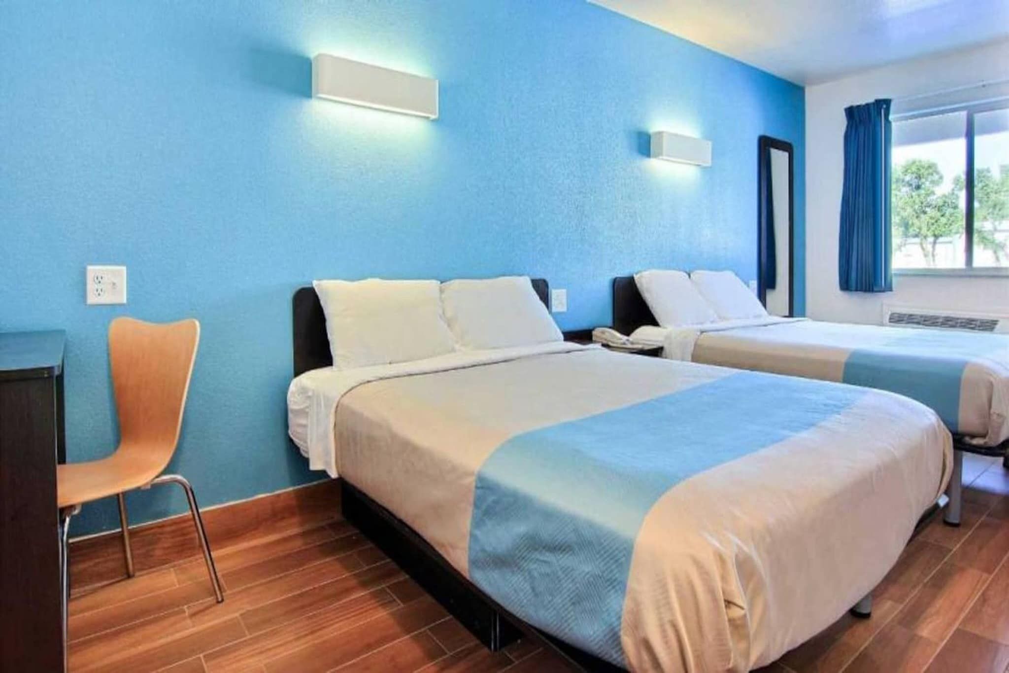 Quarto de hotel com duas camas, uma mesa, uma cadeira e uma janela com cortinas.