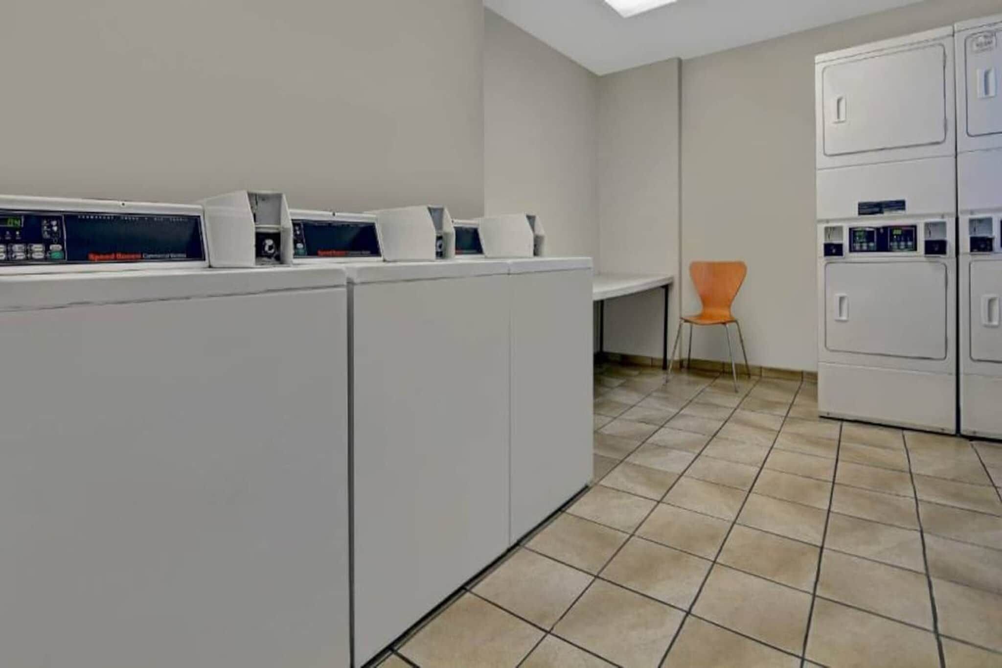 A laundry facility with multiple washing machines and dryers, a tiled floor, and a single chair.