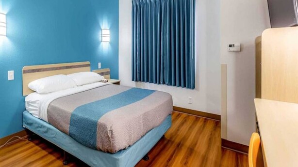 Een hotelkamer met een bed, een raam met gordijnen, een houten vloer en een stoel.