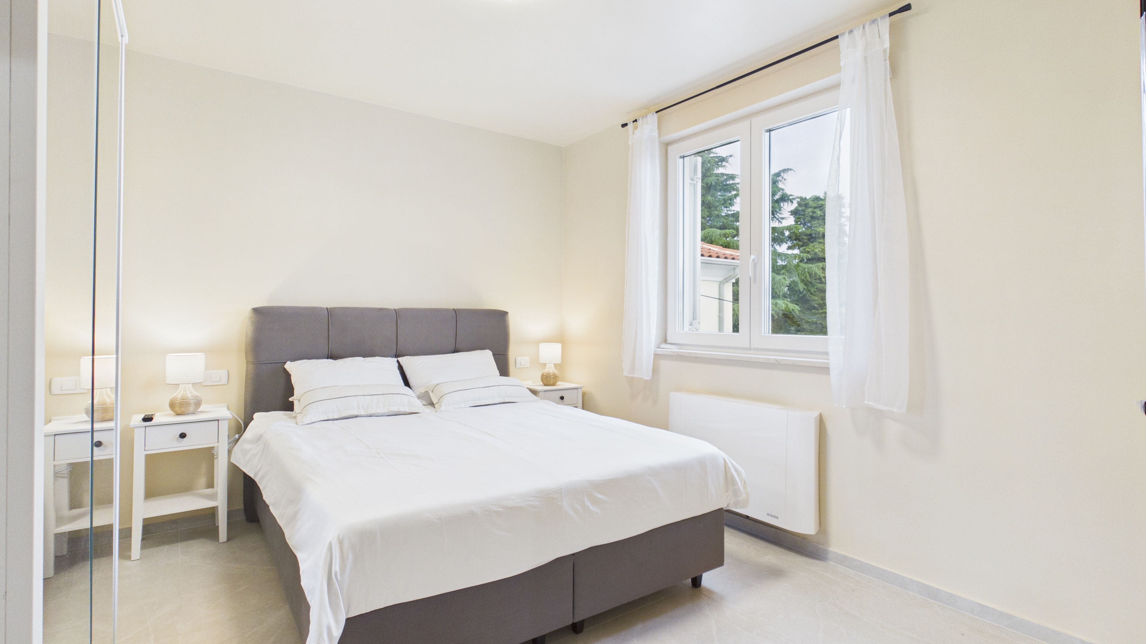 A bedroom with a bed, bedside tables, a window with curtains, and a radiator.