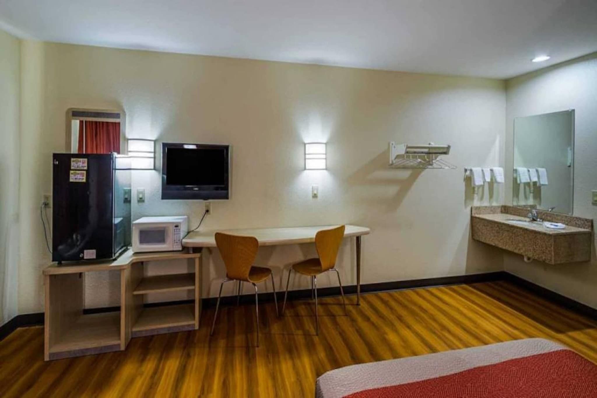 Habitación de hotel con cama, escritorio con computadora y televisor, microondas, pequeña mesa con sillas, espejo y ventana con cortinas.