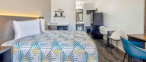 A hotel room with a bed, a desk, a chair, a kitchenette, and a bathroom.