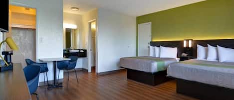 A hotel room with a wooden floor, a desk with a chair, two beds with white bedding, and a small kitchenette area.