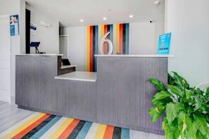 Un mostrador de recepción con el número '6' grande en la pared, una planta a la derecha y una alfombra con rayas de colores en el suelo.