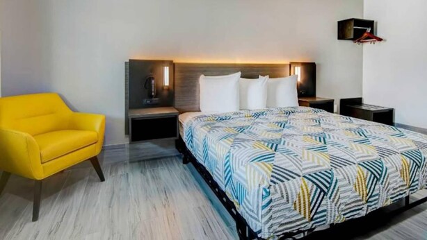 Una camera d'albergo con un letto, un comodino, una lampada, una sedia e una TV a parete.