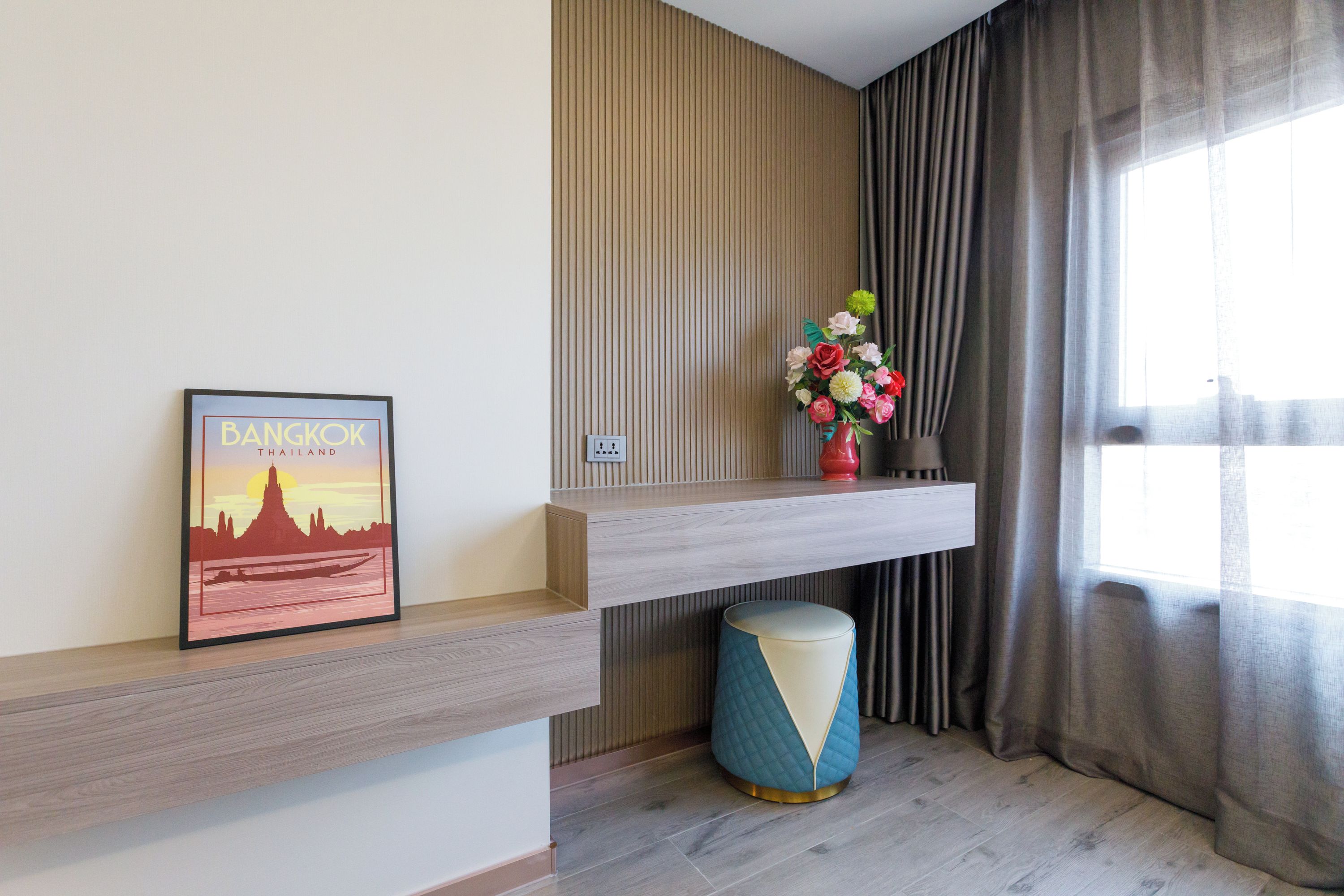 Un interior moderno con un cuadro enmarco de Bangkok, un jarrón decorativo con flores y un taburete redondo azul y blanco.