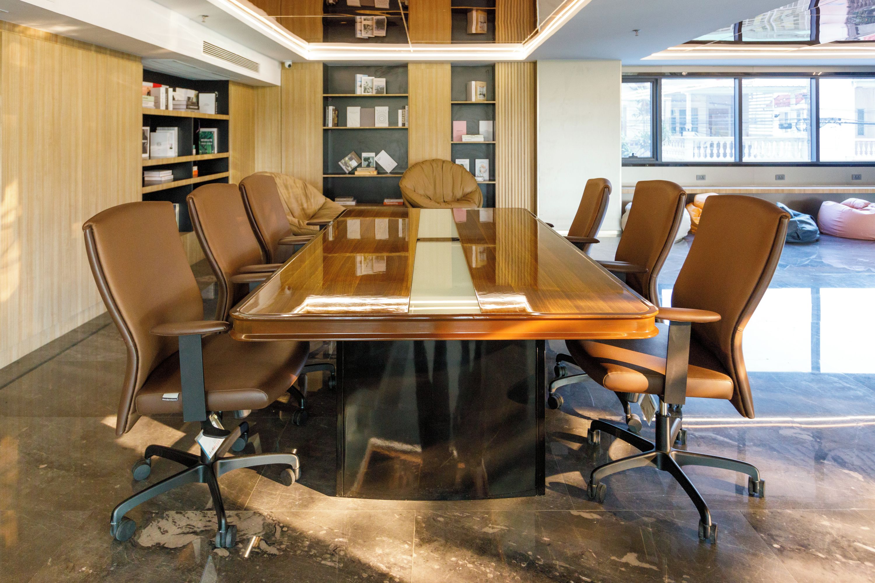 A conference room with a glass-top table surrounded by leather chairs, a wooden bookshelf, and a large window.