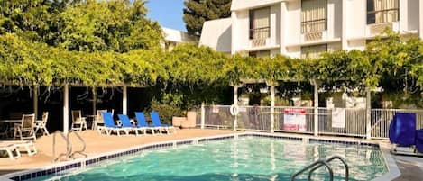 A swimming pool with blue lounge chairs and a covered area with trees.