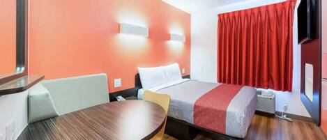 Habitación de hotel con cama, escritorio, silla, televisor y ventana con cortinas.