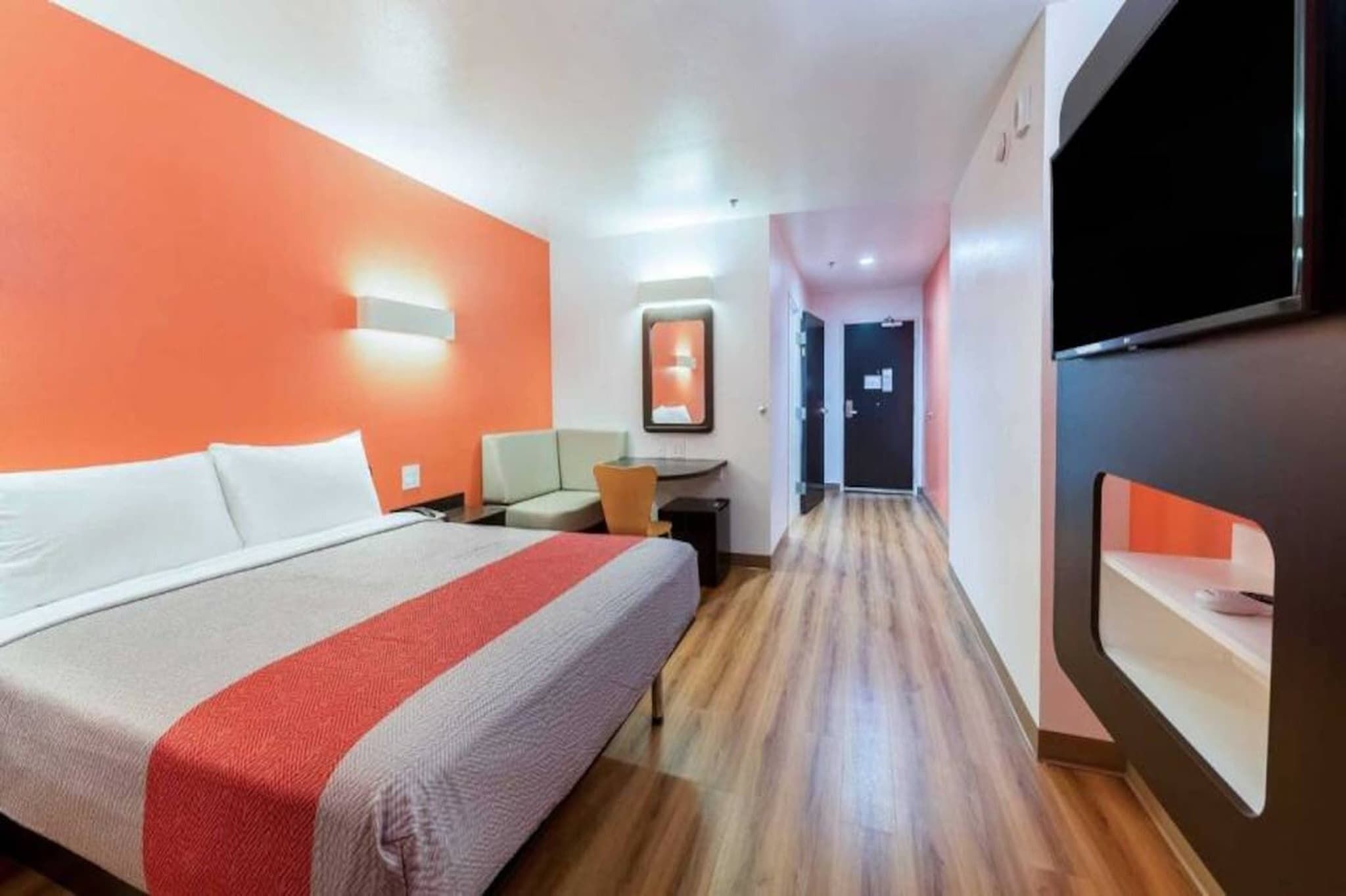 A hotel room with a bed, a desk, a chair, a television, and a hallway.