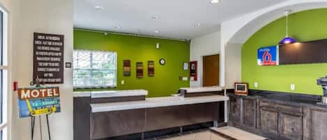 Zona de recepción de un motel con pared verde, mostrador de registro y un letrero que indica servicios de televisión y teléfono.