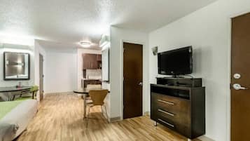 A compact room with a wooden floor, a desk with a chair, a television on a stand, and a wardrobe.