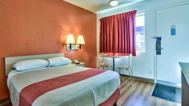 A hotel room with a bed, two bedside lamps, a desk, a chair, a window with curtains, and a door with a sign. - ADA Full Bed|Tub|Micfridge (Flaggstaff)