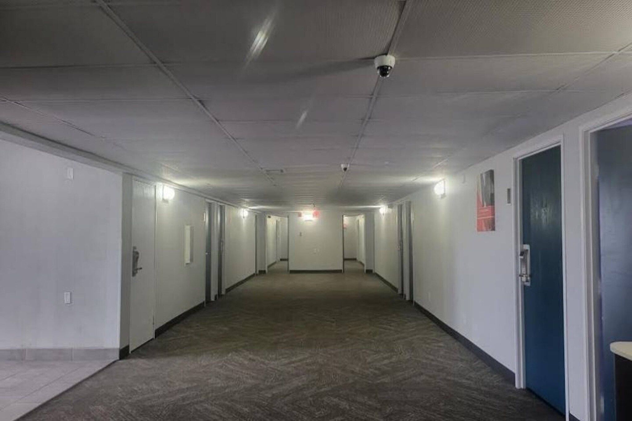 Un long couloir étroit, avec des portes de part et d’autre, un sol recouvert de moquette et un plafond où l’on peut voir les luminaires.