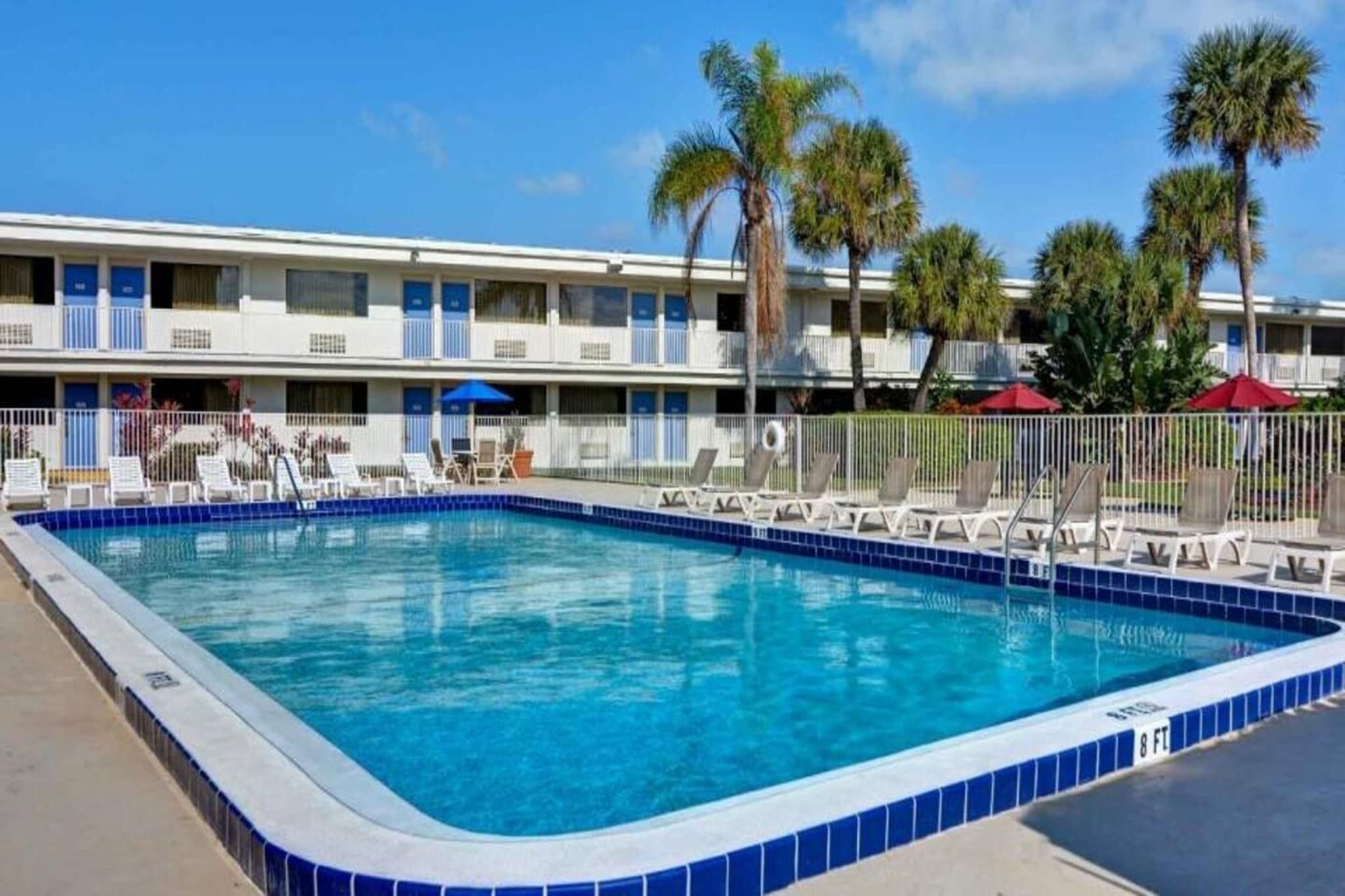 A rectangular swimming pool with a blue tiled edge, surrounded by lounge chairs and a fence.
