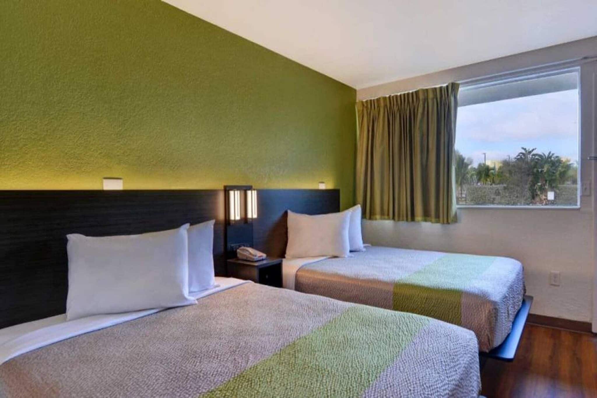 Ein Hotelzimmer mit zwei Betten, einem Nachttisch, einem Fenster mit Vorhängen und Blick auf Grün.
