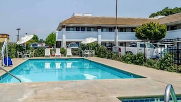 A swimming pool with lounge chairs and umbrellas, surrounded by a fence and a building with multiple windows.