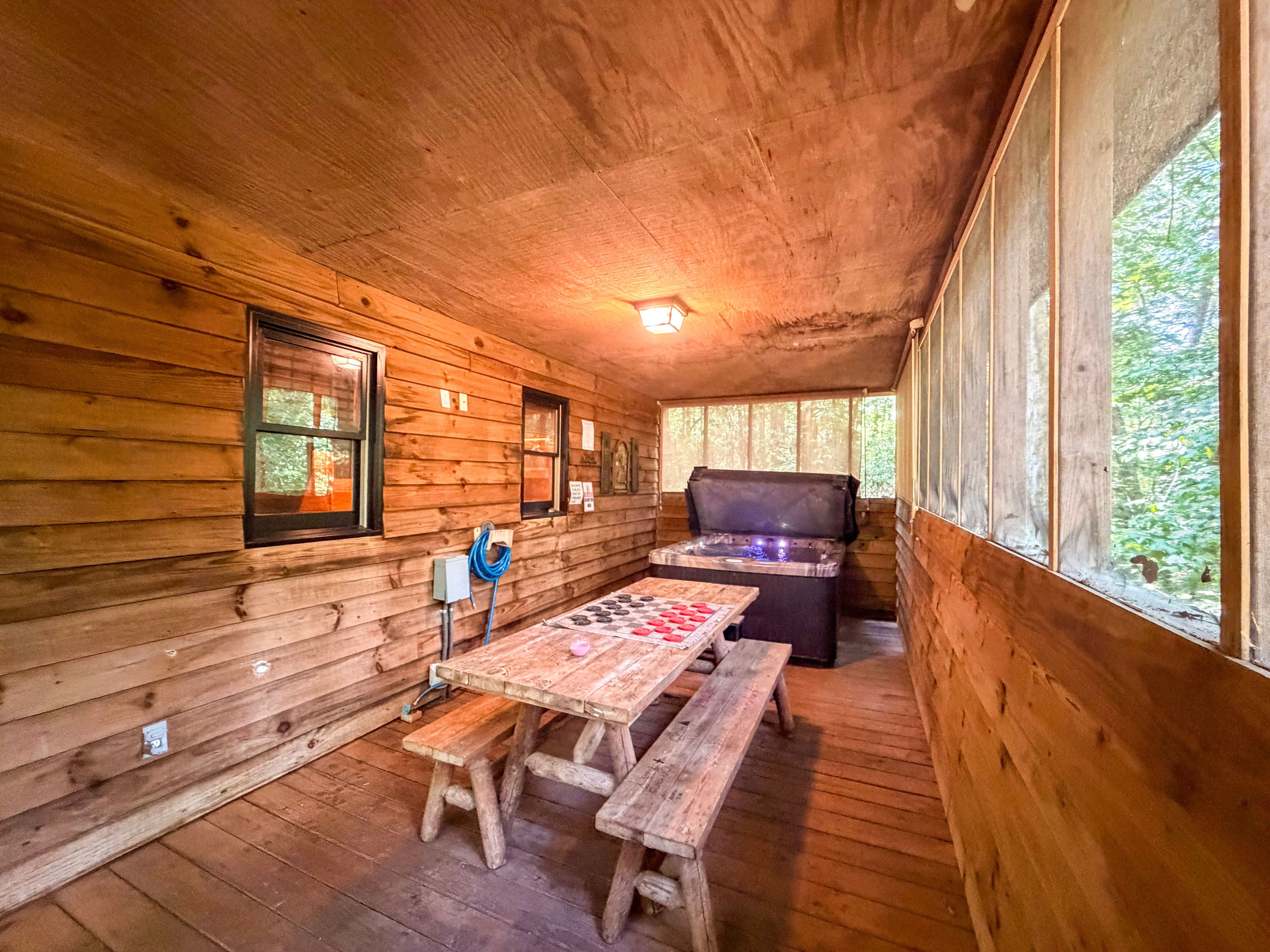 A wooden cabin interior with a wooden table and benches, a hot tub, and a window with a view of greenery.