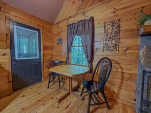 L’intérieur d’un chalet en bois comprend une table à manger, deux chaises, une fenêtre avec des rideaux, un élément de décoration fixé au mur et une cheminée en pierre.