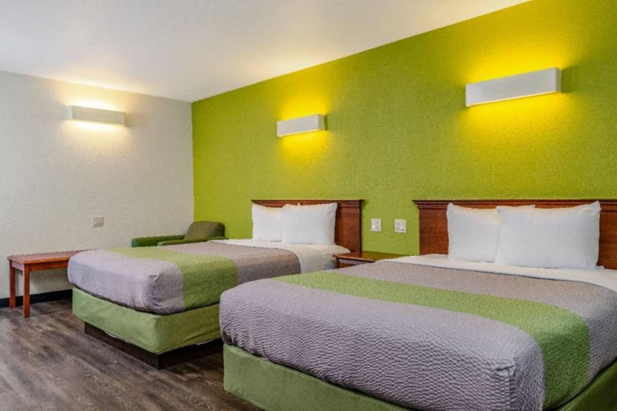 A hotel room with two beds, a small table, and a green wall with two wall-mounted lights.