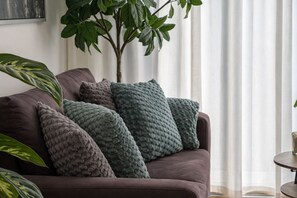 Um sofá com várias almofadas em um quarto com uma planta e uma janela com cortinas.