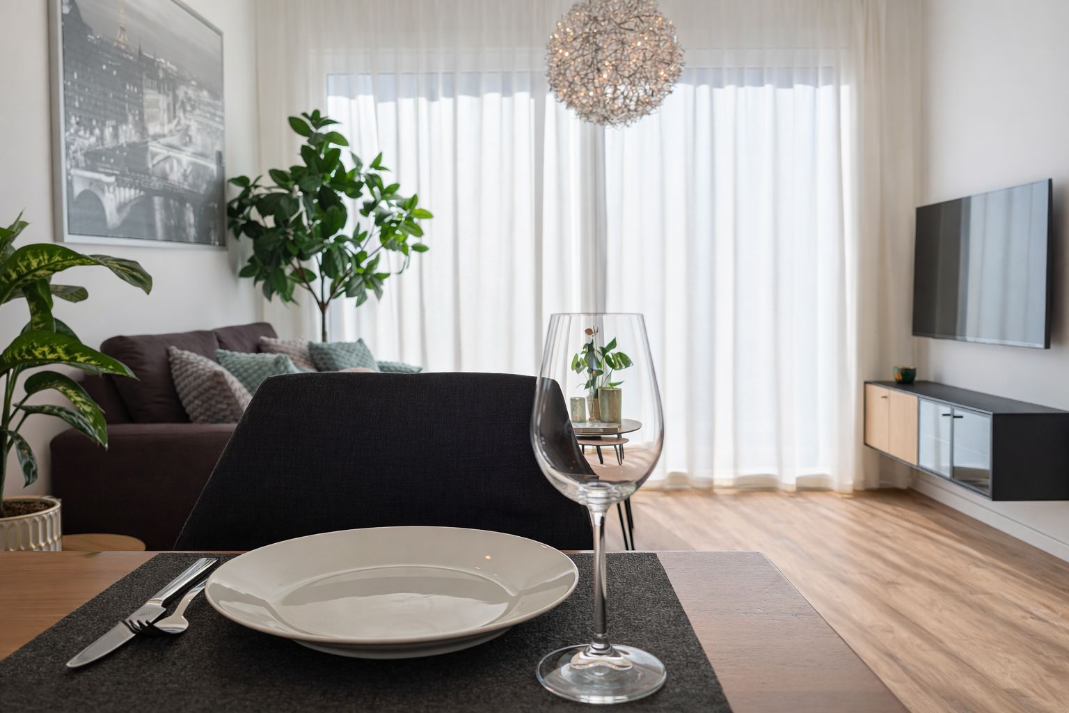 Een eetgedeelte met een tafel gedekt voor twee personen, een wijnglas en een bord. Achtergrond toont een bank en een televisie.