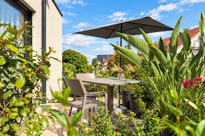 Um pátio externo com mesa e cadeiras, rodeado por vegetação exuberante e um guarda-sol grande.